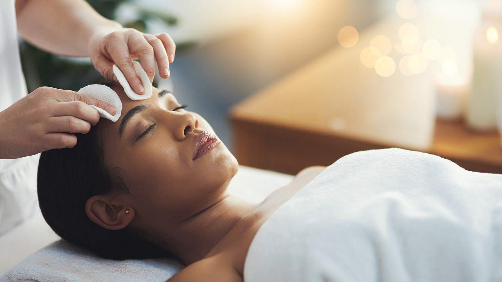 black-woman-pampered A woman lies on a massage table with her eyes closed, covered by a white towel, while a person gently cleanses her forehead with cotton pads in a calm, softly lit spa setting.