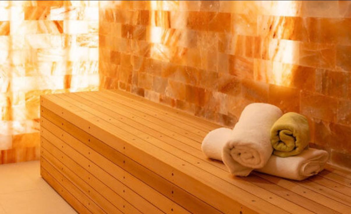 Screenshot A wooden bench with three neatly rolled towels is set against warm, glowing walls made of large Himalayan salt blocks, creating a calming and relaxing spa atmosphere.