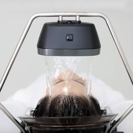 Yume Head Spa image1 A person lies back with their head under a machine washing their hair, with water flowing directly onto their scalp in a salon or spa setting.