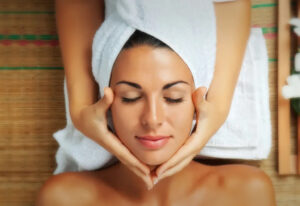 A woman with a towel wrapped around her head relaxes with her eyes closed while receiving a facial massage. She is lying on a bamboo mat, and the scene conveys a sense of calm and wellness.