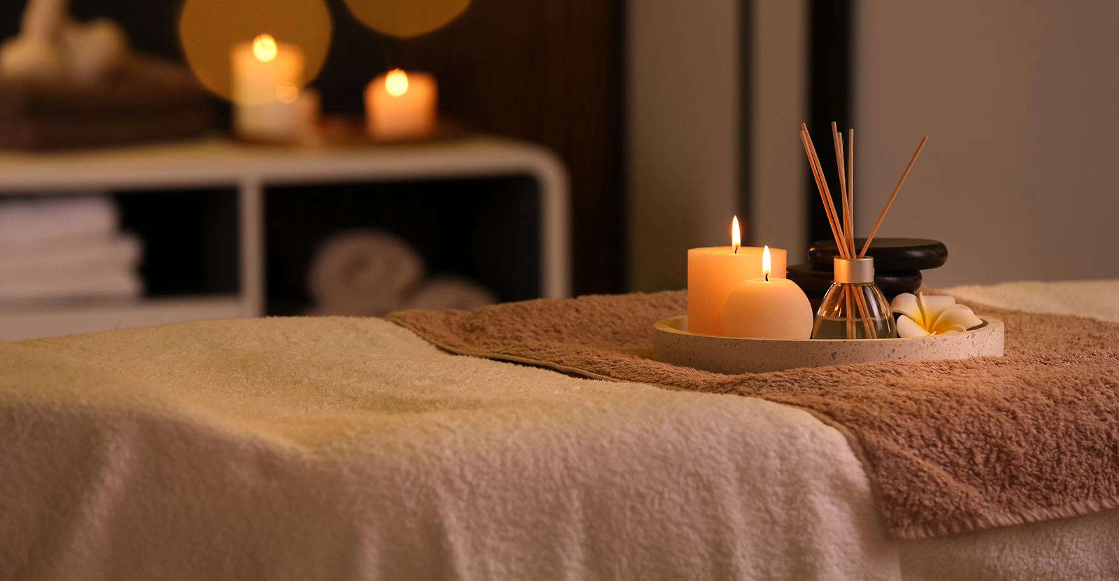 detox-table A massage table with neatly folded towels and a tray holding lit candles, aroma sticks, and spa stones, creating a warm, relaxing atmosphere with soft lighting in the background.