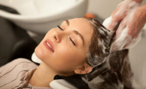 A woman with closed eyes relaxes while a hairdresser washes her hair with shampoo at a salon, her head resting in a sink, and foam visible in her hair.