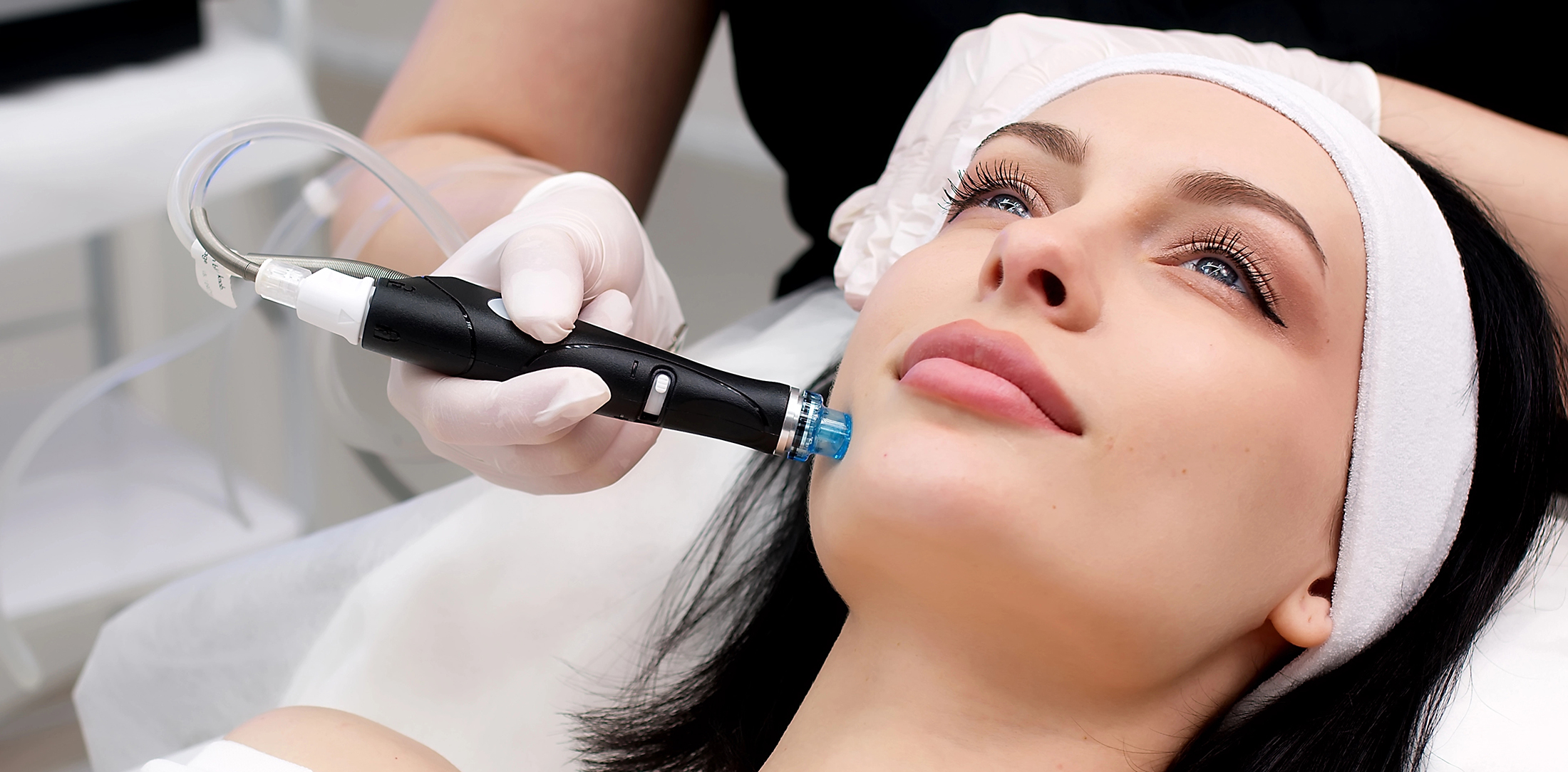 hydrafacial-24MAR A woman with a white headband receives a facial treatment from a professional using a black handheld device, while lying on a treatment bed. The setting appears to be a skincare or beauty clinic.