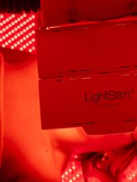 A person is lying under a LightStim ProPanel device emitting bright red LED light, typically used for light therapy or skincare treatments. The scene is bathed in a vivid red glow.