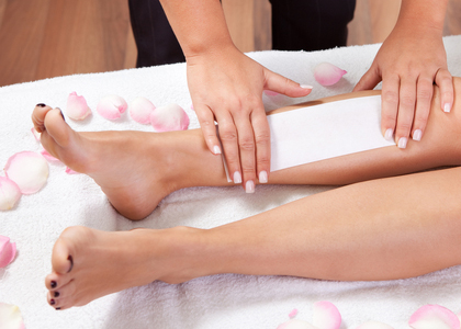 waxing-legs A person applies a waxing strip to another person’s lower leg, which is lying on a towel surrounded by scattered rose petals in a spa-like setting.