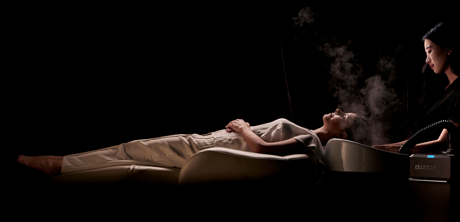 yumi-signature-spa A person lies on a reclining chair with eyes closed, receiving a facial steam treatment from an attendant in a dark room, creating a calm and relaxing atmosphere.